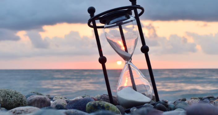 An hourglass rests on a beach with a sunset in the background